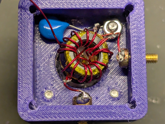 Partially-assembled unun. A nut on the upper right is connected to the coax shield, and is connected to a brass screw on the right for a counterpoise connection. A blue 100pf capacitor is in the upper left of a purple 3d-printed box, connected between ground and the center terminal of the input. At the bottom, the 6th turn is tapped and run to another brass screw which provides the 9:1 random wire connection. A strand of red magnet wire goes up to the upper left over the blue capacitor; it conn…