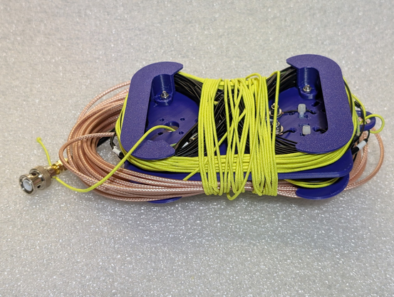Linked dipole stowed for transport. Black wire is wrapped in a figure eight on each side, with bright yellow thin elastic cord at the end of each wire. A coil of RG316 coax is underneath it, and some of the elastic cord is wrapped around the coil and winder to stow.