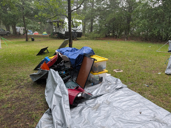 Photo of a jumbled pile of chairs, bins, and a tarp-covered table that had shortly before been covered by a pop-up shelter. In the background is an RV with a damaged awning.
